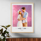 Oil-style portrait of a couple embracing on a pink and purple backdrop, in a light wood frame beside a leafy plant
