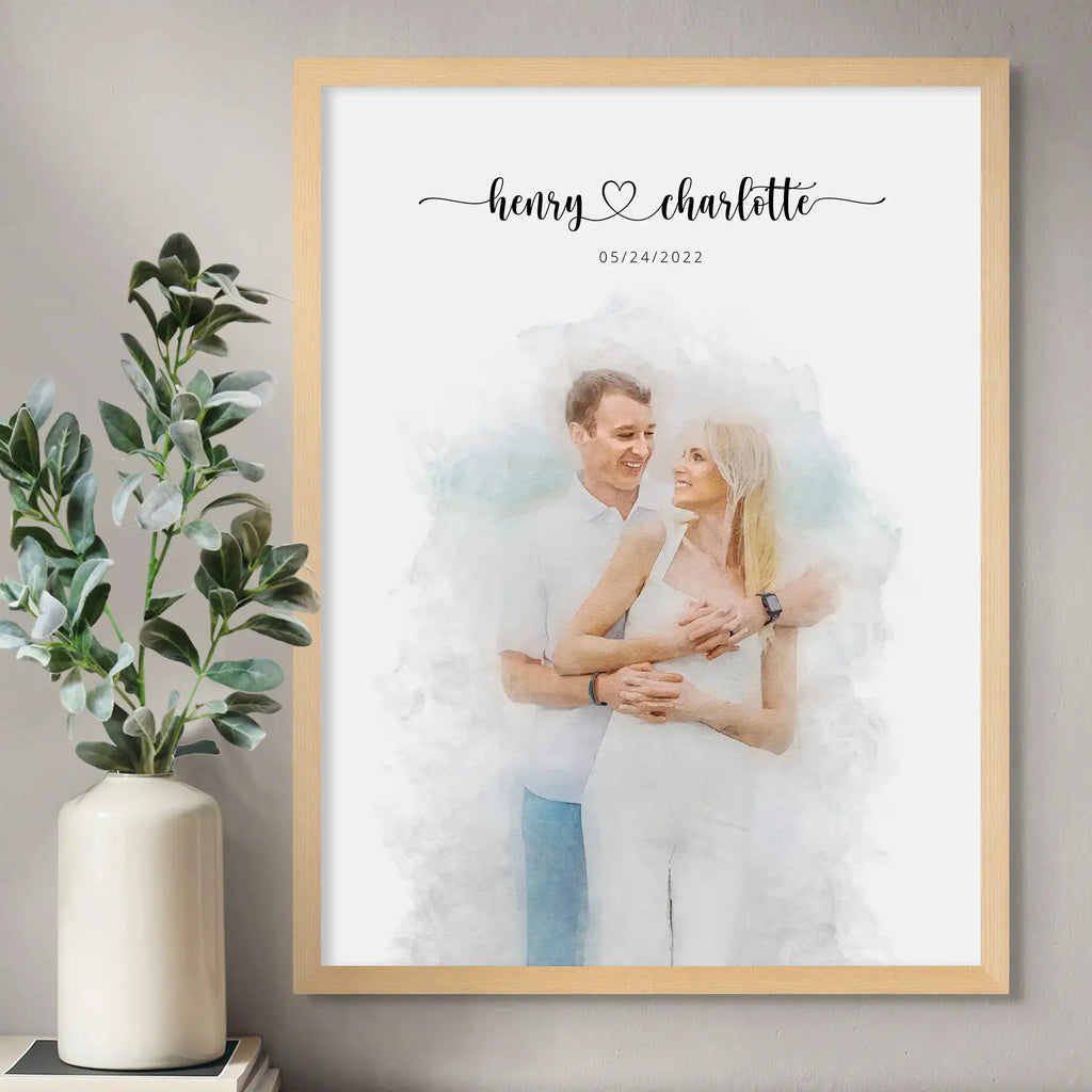Watercolor-style portrait of a smiling couple dressed in white, in a light wood frame beside a vase of leafy stems