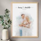 Watercolor-style portrait of a smiling couple dressed in white, in a light wood frame beside a vase of leafy stems