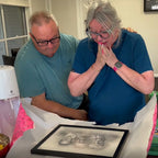 An older couple unwrap a black-framed sketch-style portrait; she presses her hands to her face