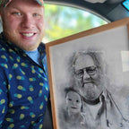 A smiling man holds up a light oak-framed sketch-style memorial portrait of a bearded man with glasses and a baby