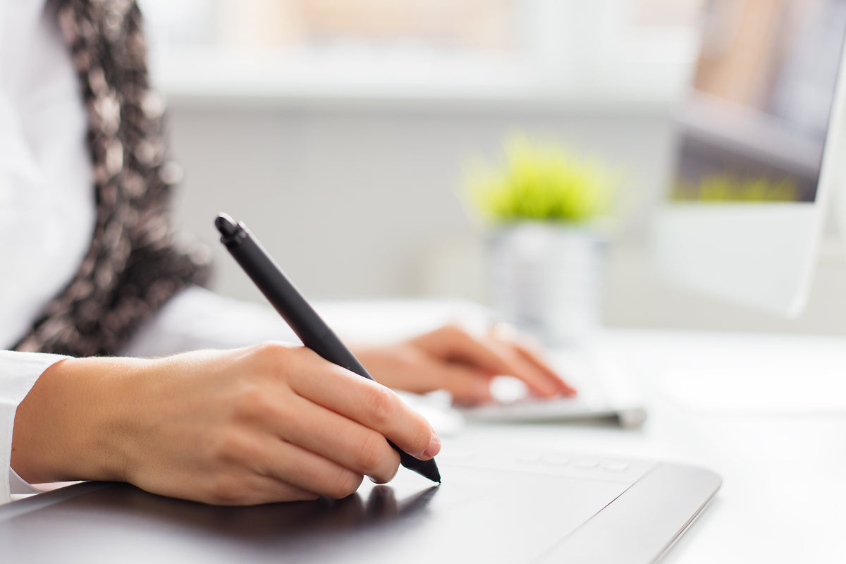 A person's hand drawing with a stylus on a graphics tablet at a desk, with a keyboard and plant behind
