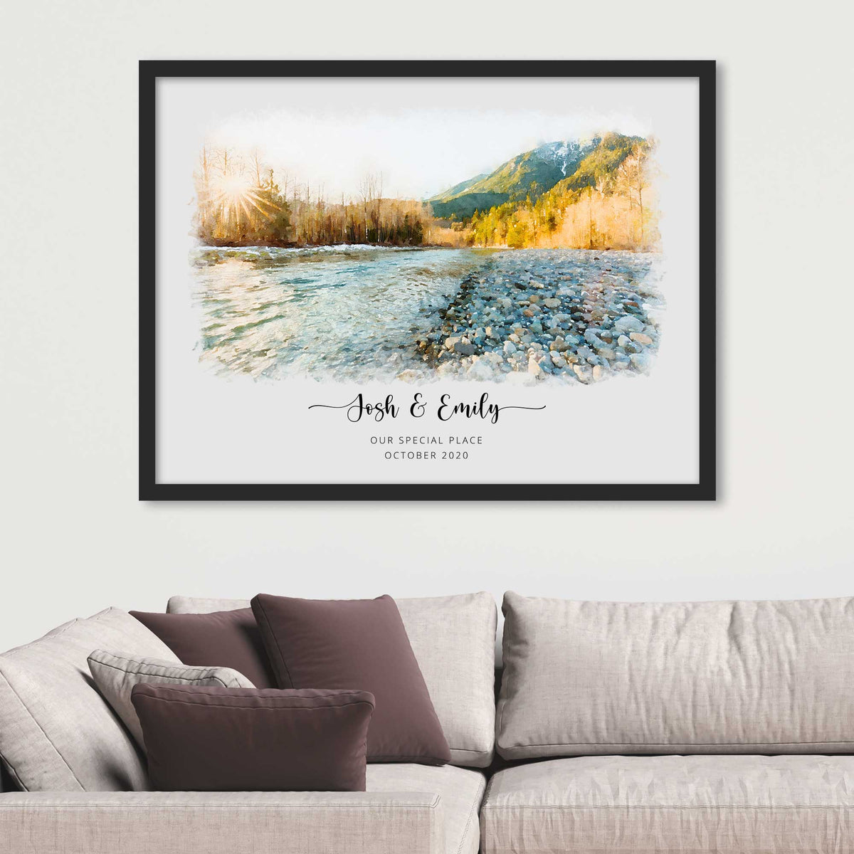 Watercolor-style portrait of a stony riverbank with sunlit trees and hills, framed in black above a sofa, names beneath