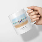 White mug held in one hand, printed with a watercolor-style portrait of a family holding hands on a beach